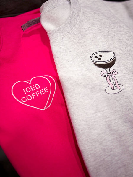 Two sweatshirts, one pink with 'ICED COFFEE' text and one gray with a cocktail graphic, on a dark surface.