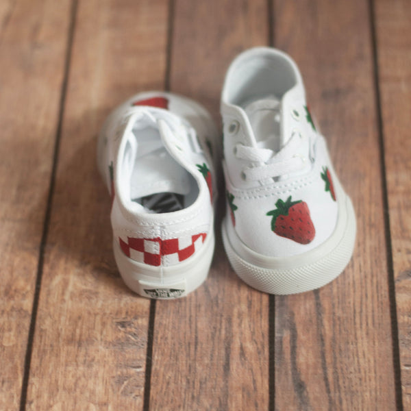 Pair of white vans sneakers with strawberry designs hand painted on the shoe sitting on a wooden floor