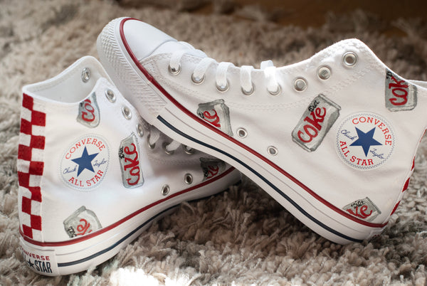 A pair of white high-top Converse All Star sneakers decorated with red and silver Coca-Cola themed patches, a red-and-white checkered heel stripe, and a blue star logo on the side.