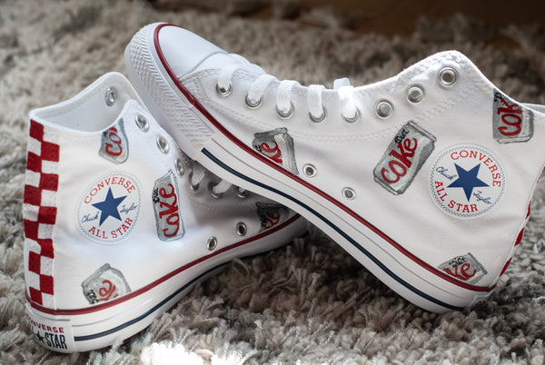 A pair of white high-top Converse All Star sneakers with custom Coca-Cola themed graphics, including red and white checkers, silver can designs, and blue star logos, resting on a gray shag rug.