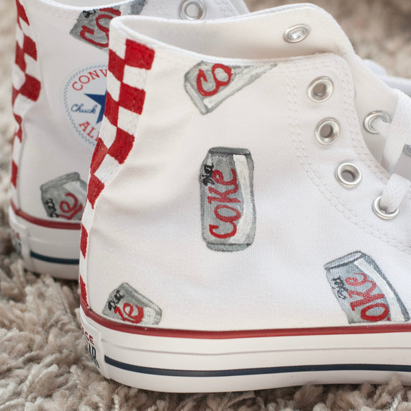 A pair of white high-top canvas sneakers with vintage-style Coca-Cola can graphics and red-and-white striped side panels, resting on a shaggy beige rug.