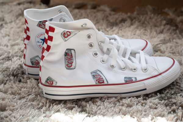 A pair of white high-top canvas sneakers with red and white checkered side panels and multiple vintage-style Coca-Cola can graphics, displayed on a shaggy gray rug.