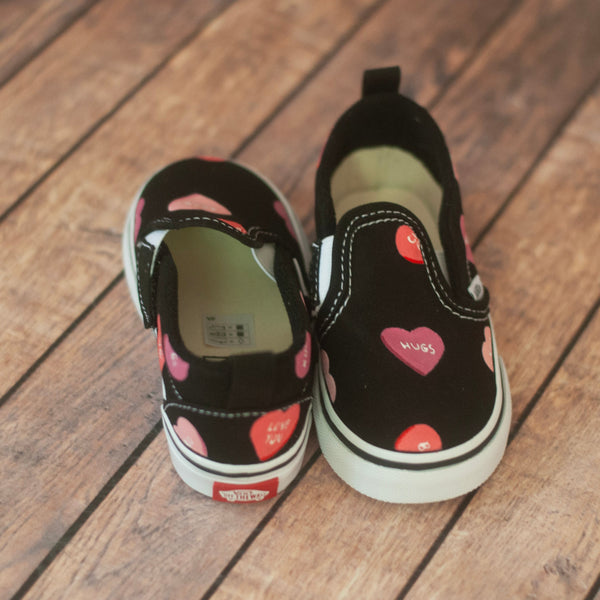 A pair of black slip-on shoes with pink and red heart patterns, displayed on a wooden plank floor.