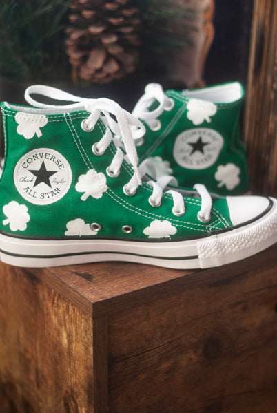 Green Converse All Star high-top sneakers with white shamrocks painted on the shoe while they sit on a wooden surface.