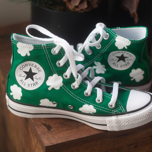 Green Converse All Star high-top sneakers with white shamrocks painted on the shoe while they sit on a wooden surface.