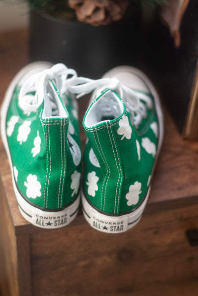 Green Converse All Star high-top sneakers with white shamrocks painted on the shoe while they sit on a wooden surface.