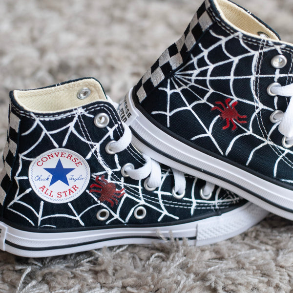 A pair of black high-top sneakers with a spider-themed design, featuring white spiderwebs and red spider silhouettes, styled on a gray shag rug.