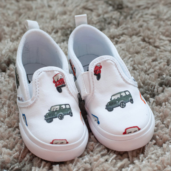 A pair of white slip-on baby shoes painted with small colorful cars resting on a beige shag rug.