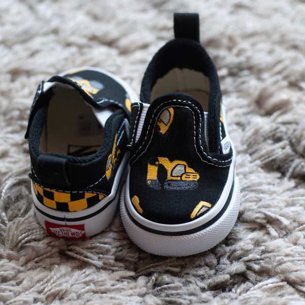 A pair of black slip-on sneakers with yellow and black checkered details with construction trucks painted on, resting on a textured gray shag rug.