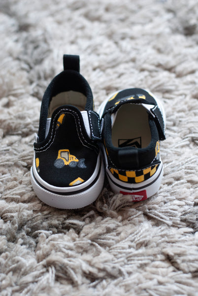 A pair of black slip-on sneakers with yellow and black checkered details with construction trucks painted on, resting on a textured gray shag rug.