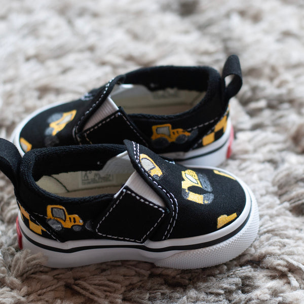 A pair of black slip-on sneakers with yellow and black checkered details with construction trucks painted on, resting on a textured gray shag rug.