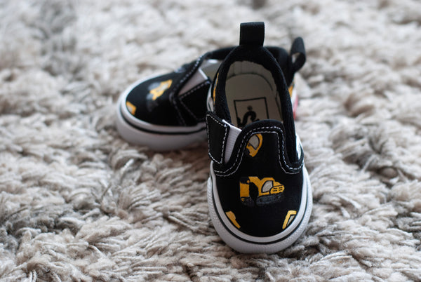 A pair of black slip-on sneakers with yellow and black checkered details with construction trucks painted on, resting on a textured gray shag rug.