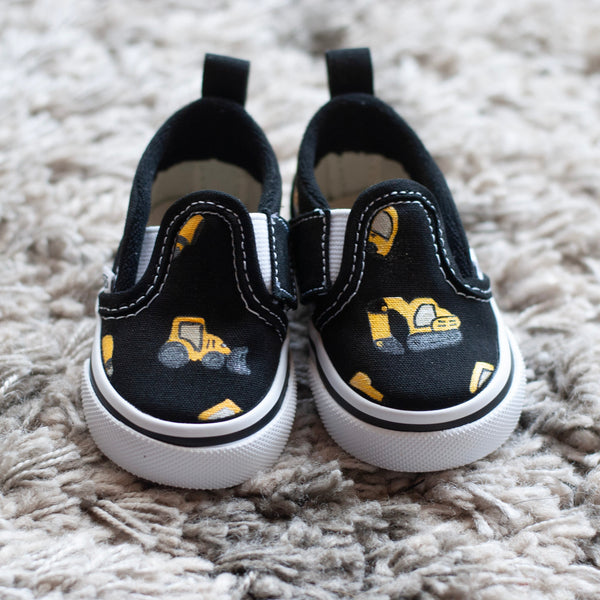 A pair of black slip-on sneakers with yellow and black checkered details with construction trucks painted on, resting on a textured gray shag rug.
