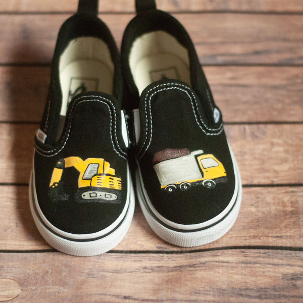 Black slip-on shoes with truck and bulldozer designs on a wooden surface
