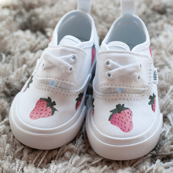 A pair of white canvas low-top sneakers with embroidered pink strawberries on the sides, resting on a textured beige shag rug.