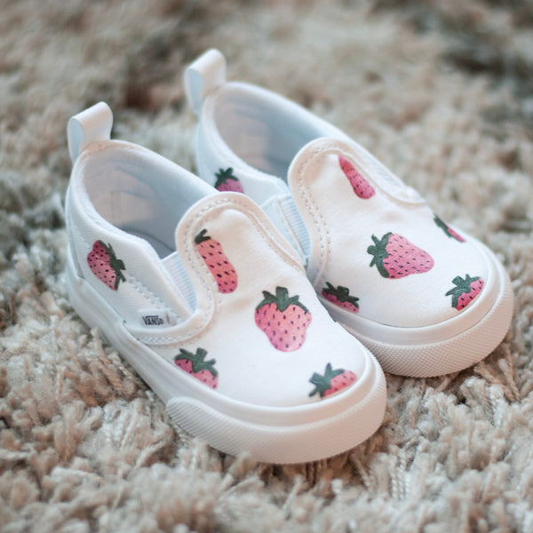 A pair of white slip-on baby sneakers with a pink strawberry pattern, resting on a textured beige shag rug.