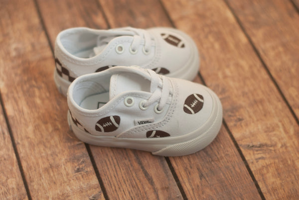 White tie Vans with brown football designs on a wooden floor. The shoe are facing sideways.