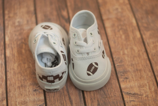 Pair of white vans, one facing forward and one facing backwards with footballs hand painted on them, scattered along the shoe. The back of the shoe has a brown checkered pattern. 