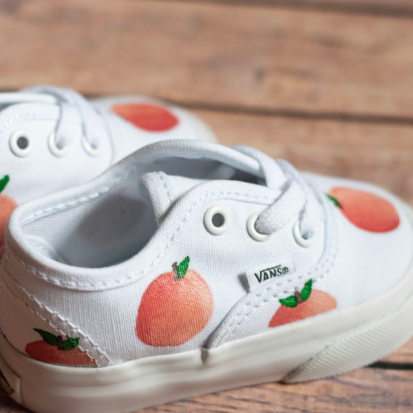 A pair of white canvas toddler shoes with hand-painted peach designs, featuring laces and the brand 'Vans' on the side.