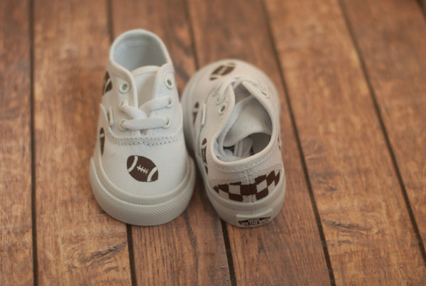 White tie toddler vans with brown footballs handpainted and scattered along the shoe on a wooden floor. The back of the shoe has a brown checkered pattern painted on. One shoe is facing forward and one is facing backwards.