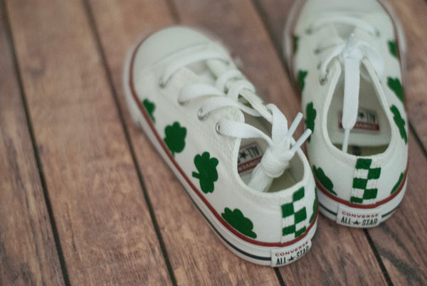 A pair of white Converse low-top sneakers with green checkered patterns on the back and shamrocks along the side on a wooden floor.