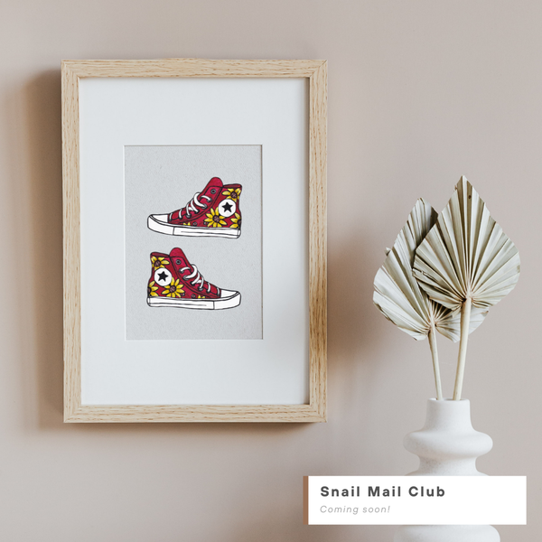 Framed artwork of red high-top sneakers with floral sunflower patterns on a beige wall, next to a decorative plant.